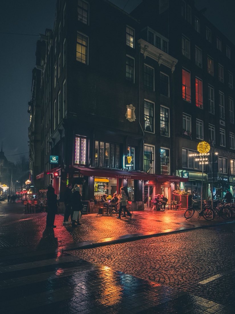 A corner bar in Amsterdam at night, warm windows and rain on the cobbles