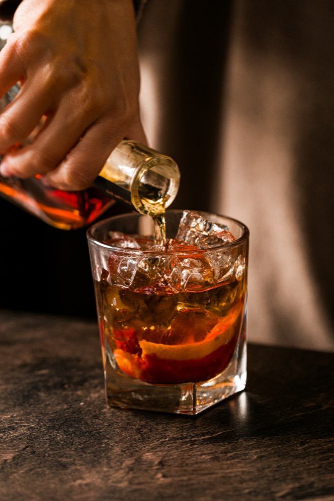 A bartender pouring amber spirit over ice in a heavy tumbler