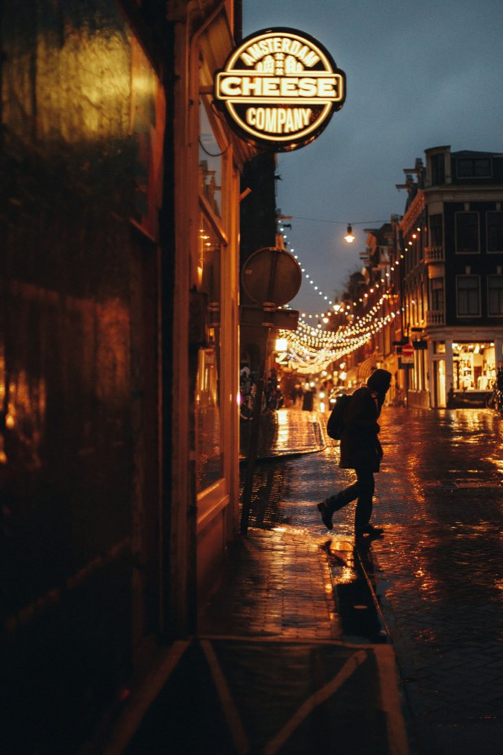 A lone figure crossing a wet Amsterdam street at night under strings of lights